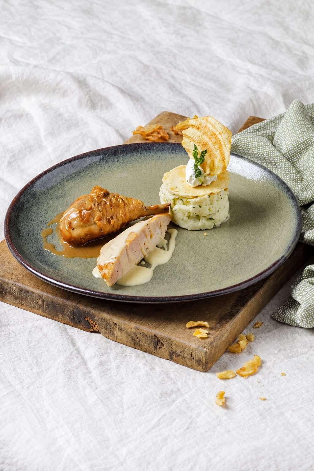 Une assiette en céramique grise contient une cuisse de volaille laquée, un morceau de blanc avec sa sauce crémeuse et une timbale de purée surmontée d'une chips, posée sur une planche en bois.