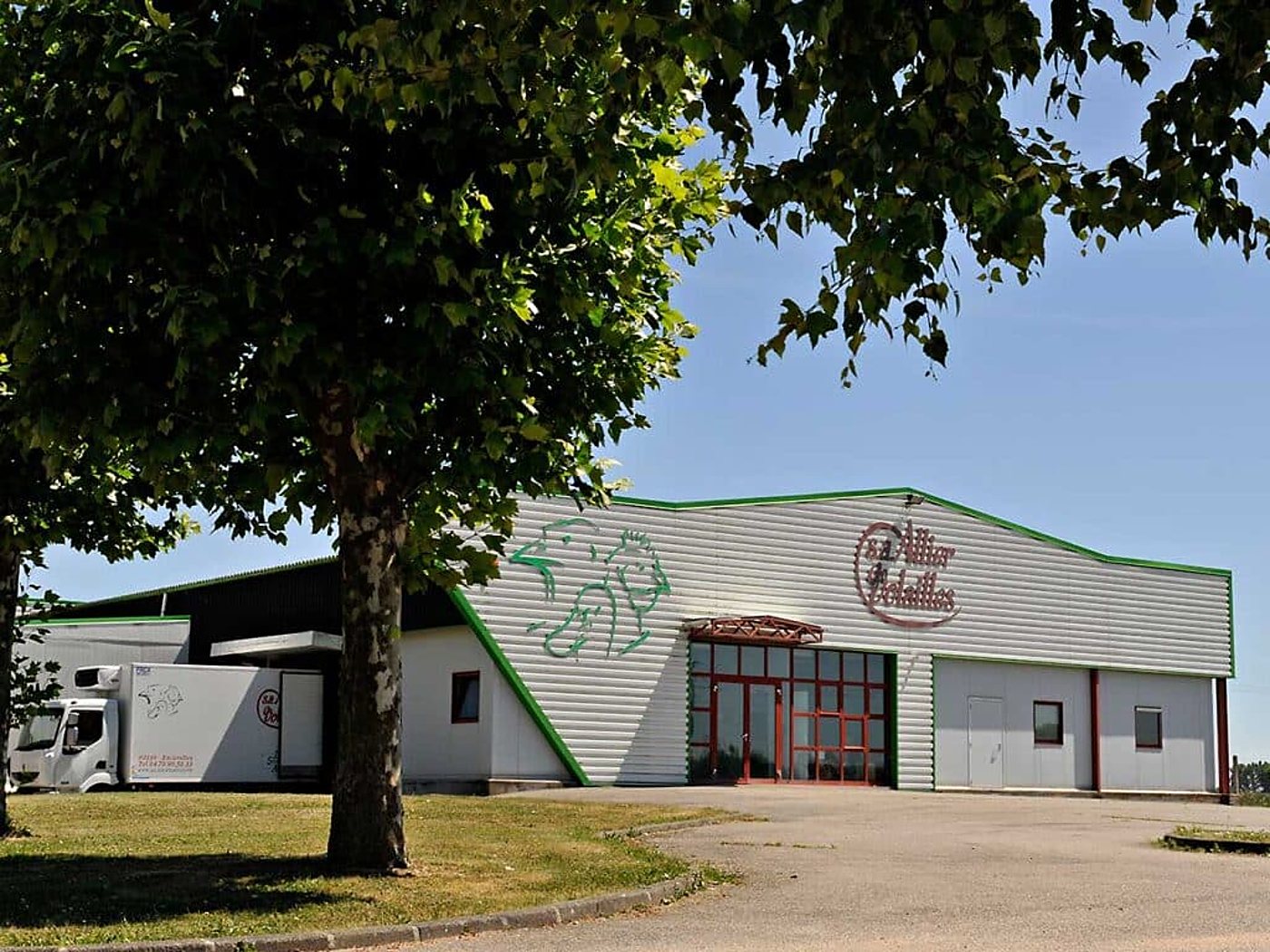 Façade d'un entrepôt blanc à liseré vert portant l'enseigne Allier Volailles, avec un camion frigorifique garé à gauche sous des arbres.