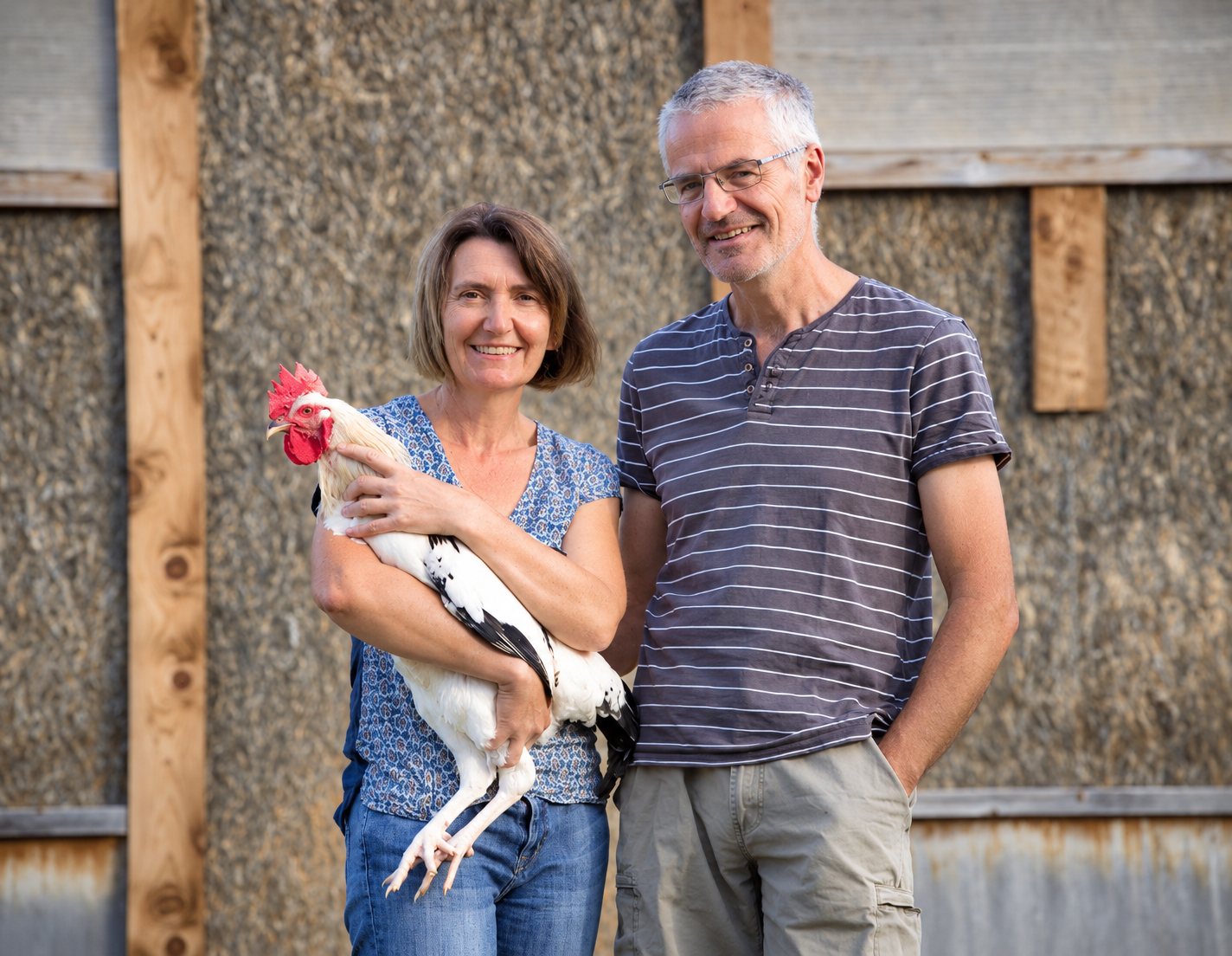 Une femme et un homme souriants posent côte à côte devant un mur de grange en bois, la femme tenant un coq blanc à crête rouge dans ses bras.
