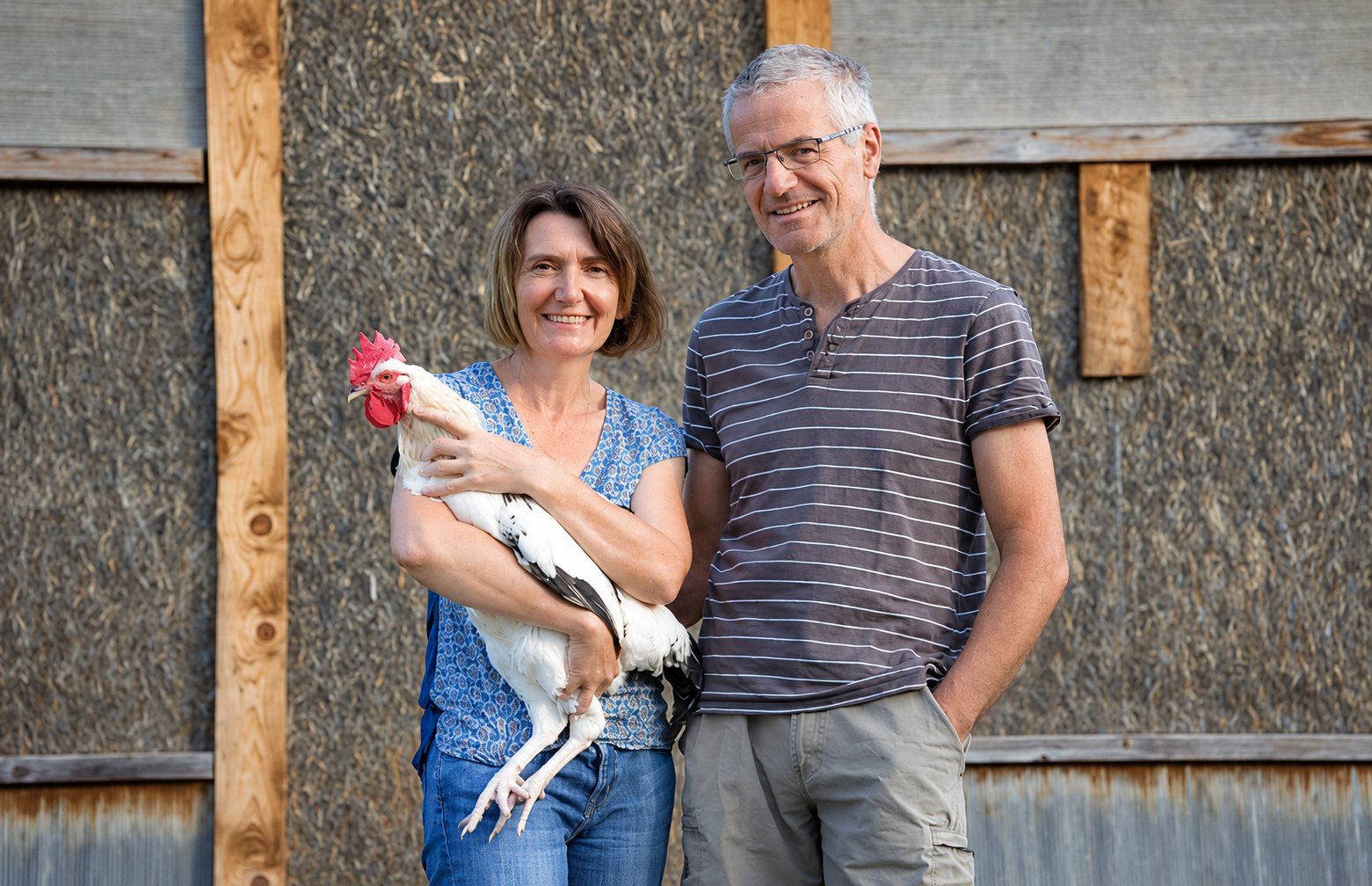 Une femme et un homme souriants posent devant un mur de paille et de bois, la femme tenant un coq blanc dans ses bras.