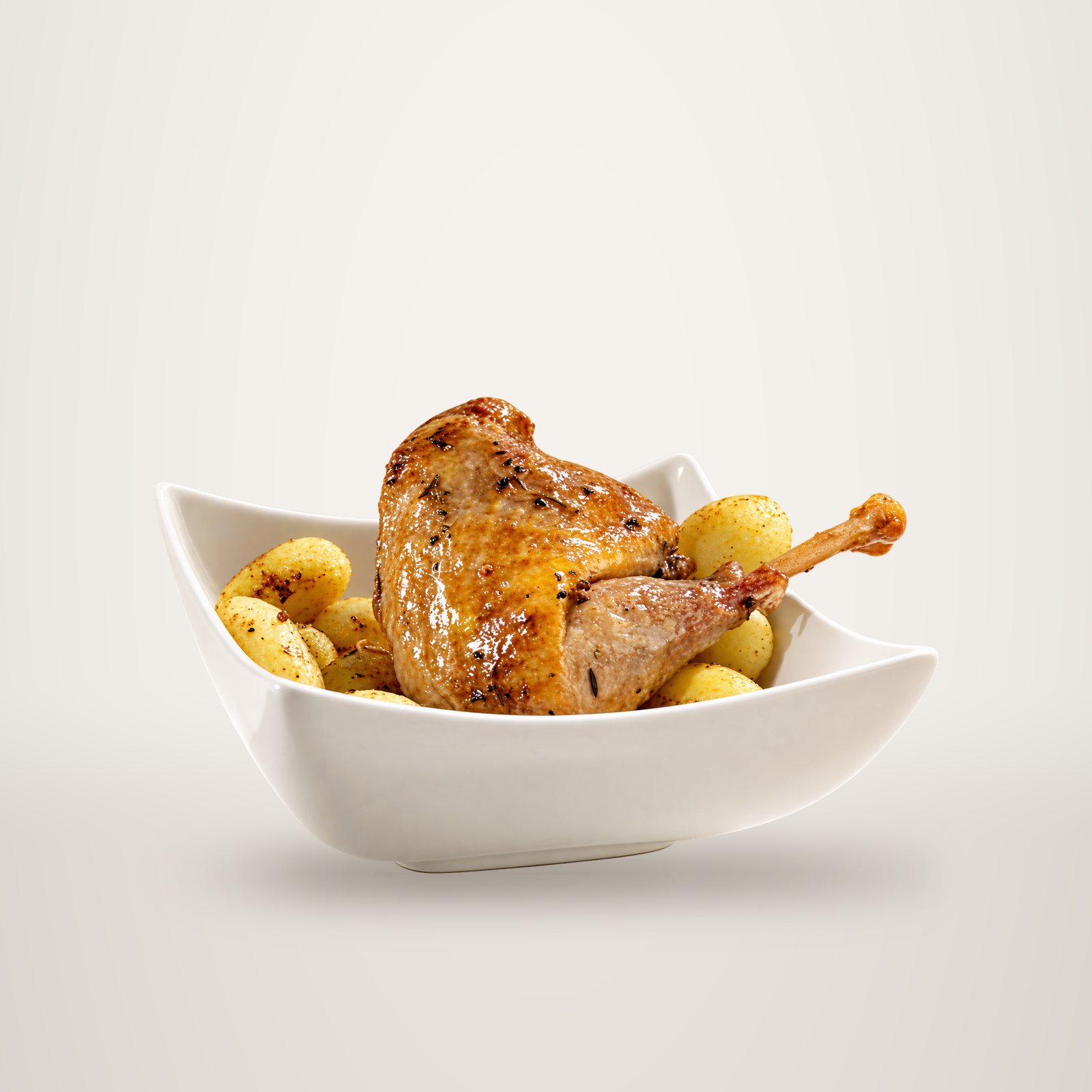 Une cuisse de volaille rôtie et dorée, parsemée d'herbes et de poivre, servie sur des pommes de terre sautées dans un bol blanc carré.