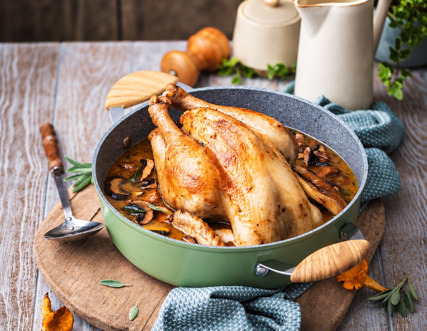Un poulet rôti doré avec une sauce aux champignons dans une poêle verte à poignée en bois, posée sur une planche ronde et une table en bois, avec une louche et des herbes autour.