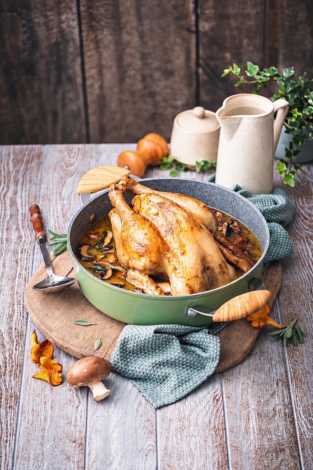 Un poulet rôti avec des champignons et une sauce dorée dans une poêle verte posée sur une planche en bois, avec un torchon, une cuillère et une cruche beige en arrière-plan.