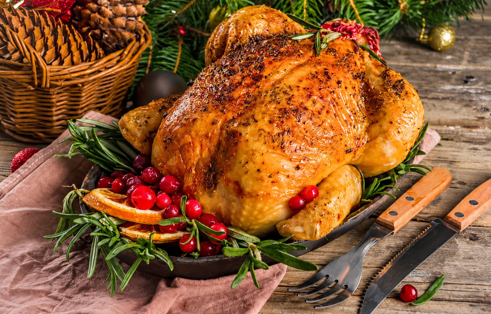 Un poulet rôti doré servi sur un plat avec du romarin, des canneberges et des tranches d'orange, près d'un panier de pommes de pin et de branches de sapin sur une table en bois.