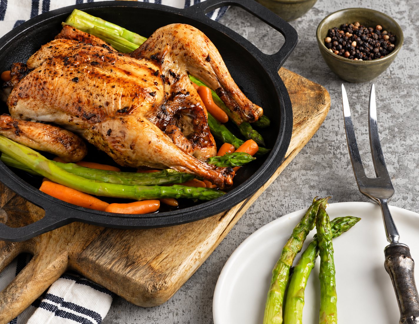 Un poulet entier rôti dans une poêle en fonte noire, entouré d'asperges et de carottes, posée sur une planche en bois avec une assiette d'asperges, une fourchette à découper et un bol de poivre en grains.
