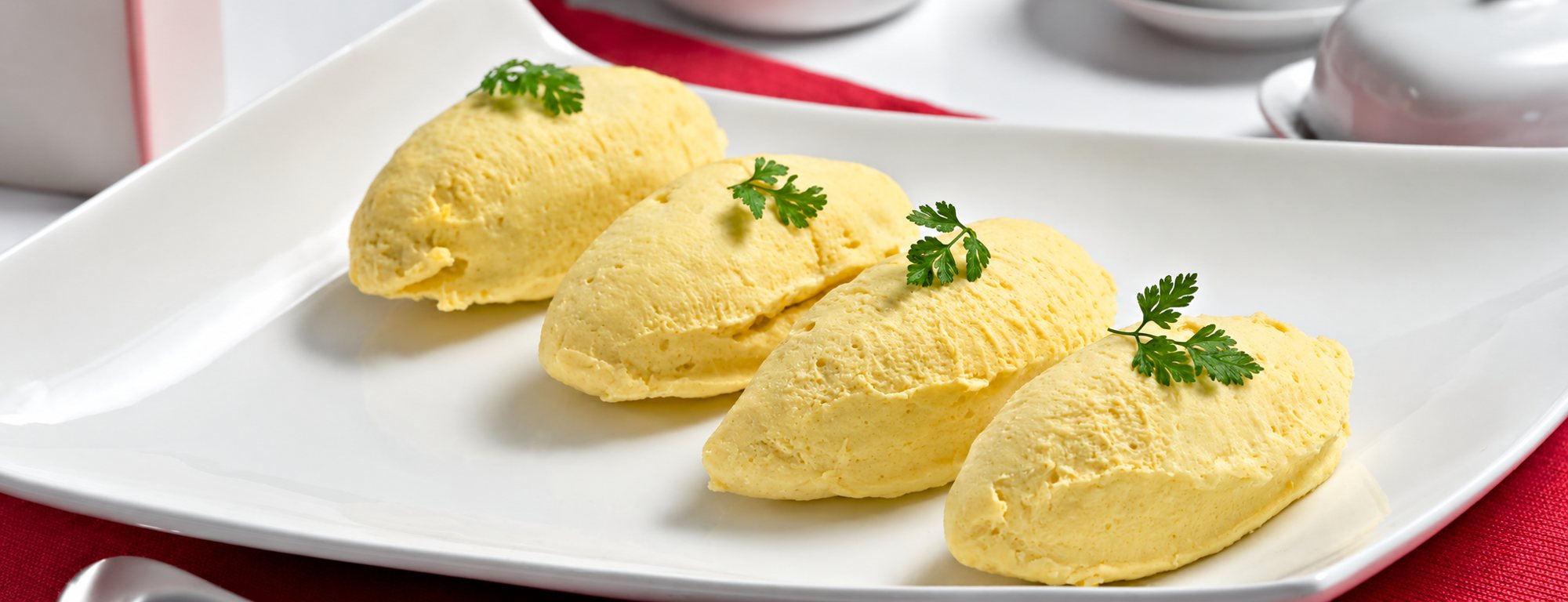 Quatre quenelles de mousse jaune pâle décorées d'une feuille de cerfeuil, présentées sur une assiette rectangulaire blanche posée sur une nappe rouge.