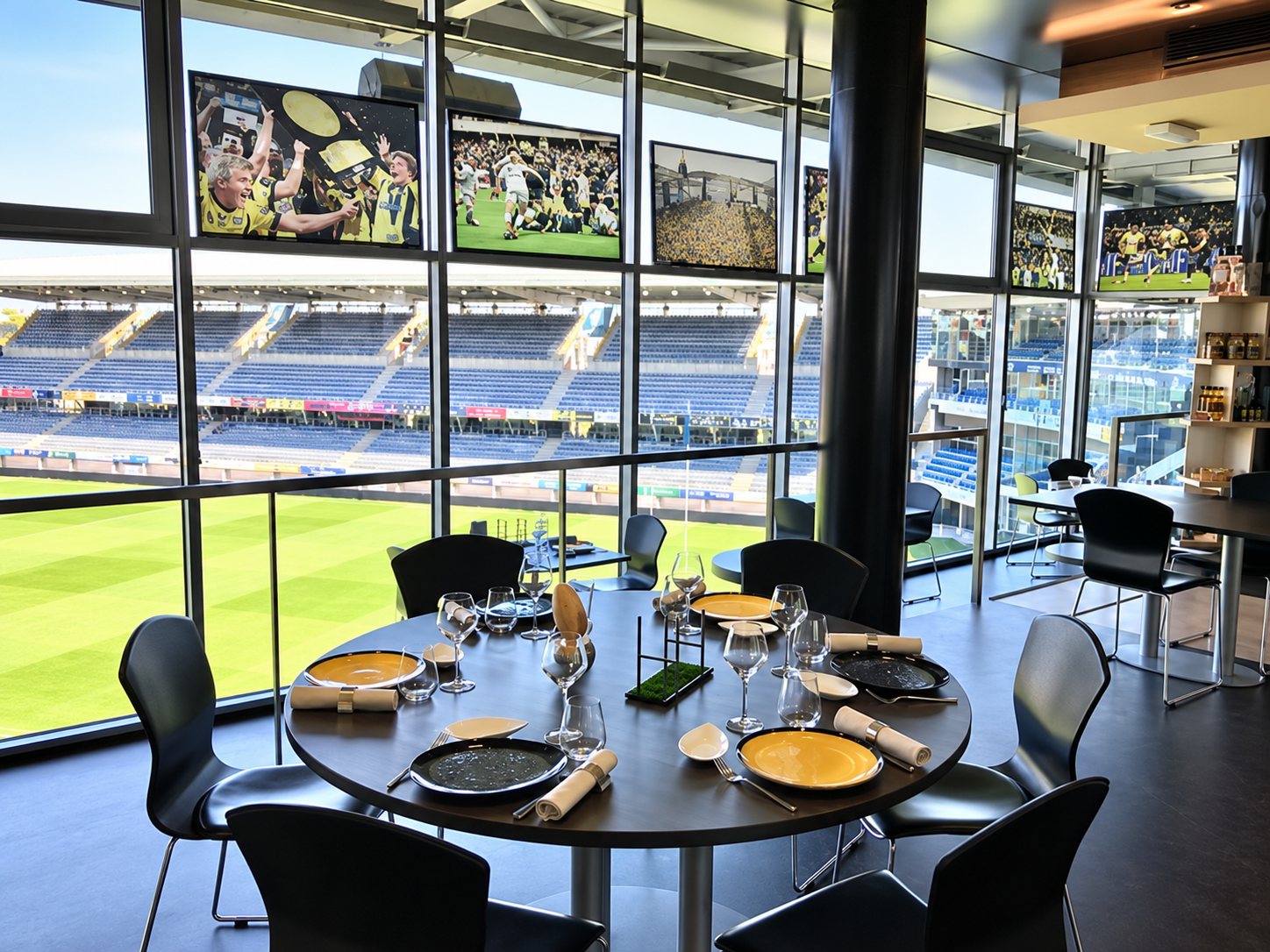Une table ronde élégamment dressée avec assiettes jaunes et verres se trouve dans une salle de restaurant dont les grandes baies vitrées donnent sur un stade et sa pelouse.