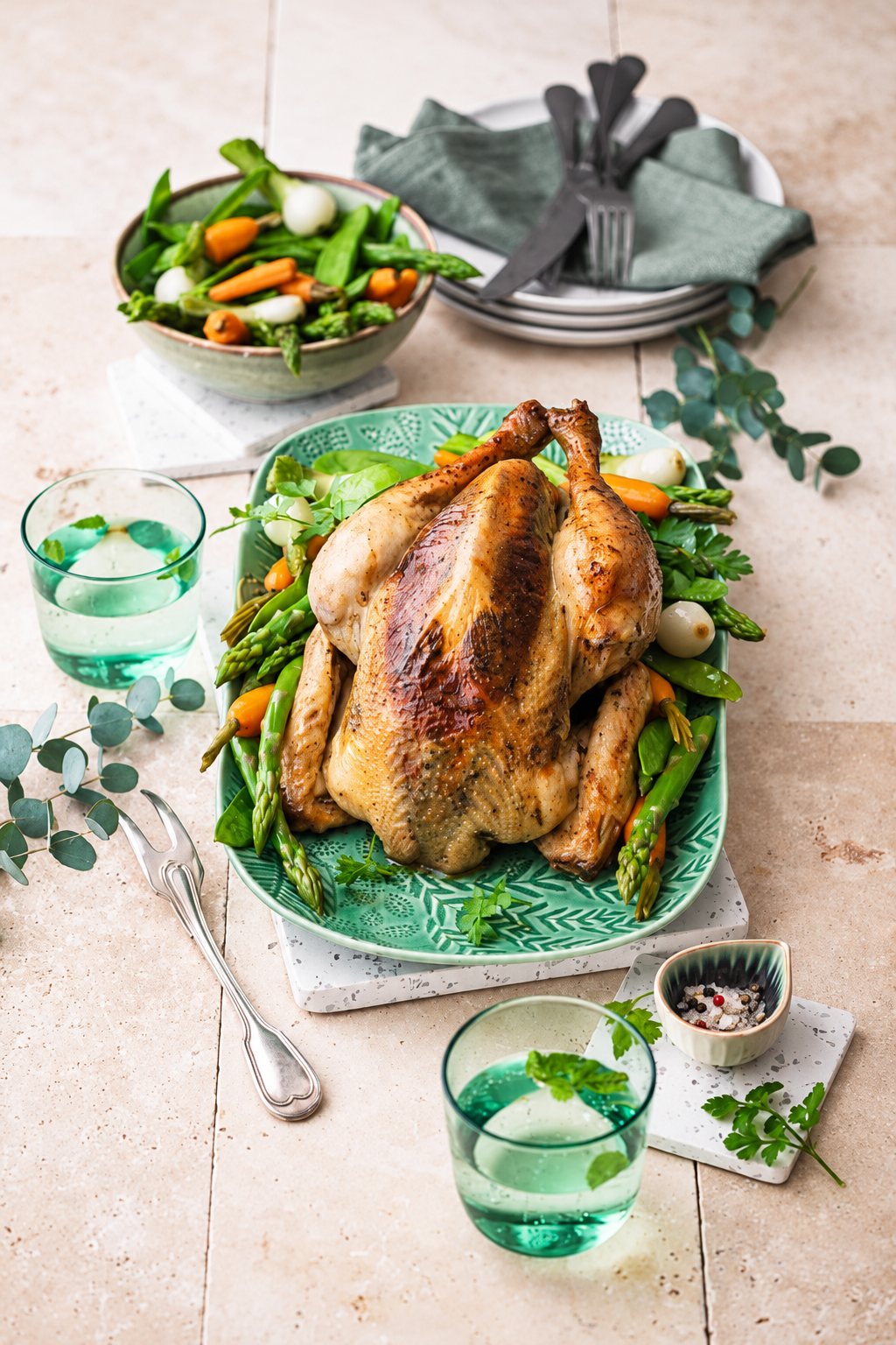 Une volaille entière rôtie est servie sur un plat vert avec des asperges, des carottes et des oignons nouveaux, accompagnée de verres d'eau et d'un bol de légumes sur une table en carreaux beiges.